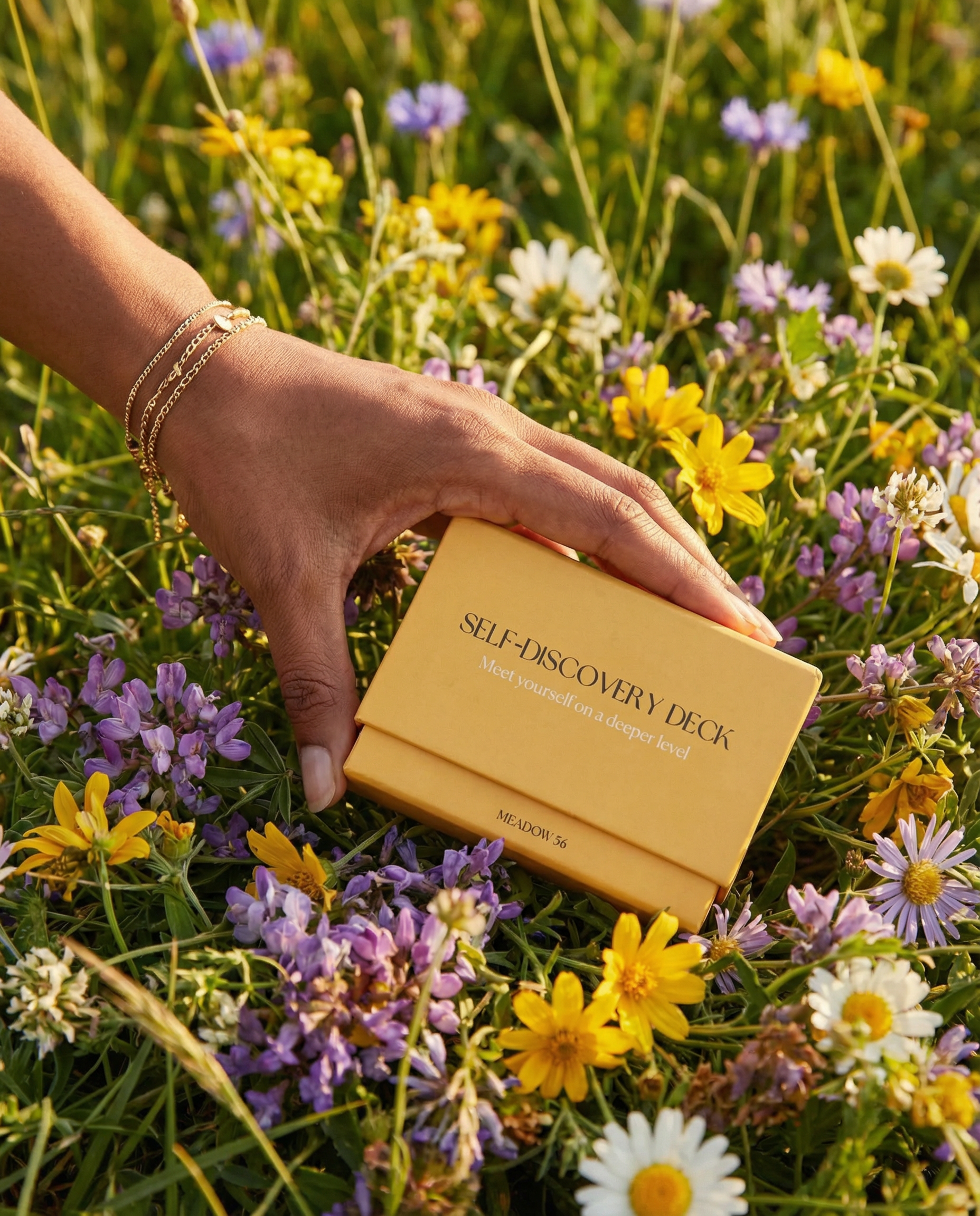 Hand holding a 'Self-Discovery Deck' box among wildflowers